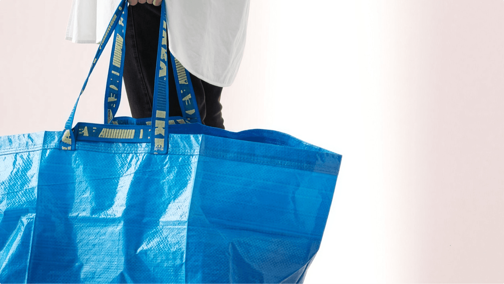 A person carries IKEA's large blue FRAKTA tote bag, its woven plastic body and yellow-lettered handles filling the frame against a soft pink background.