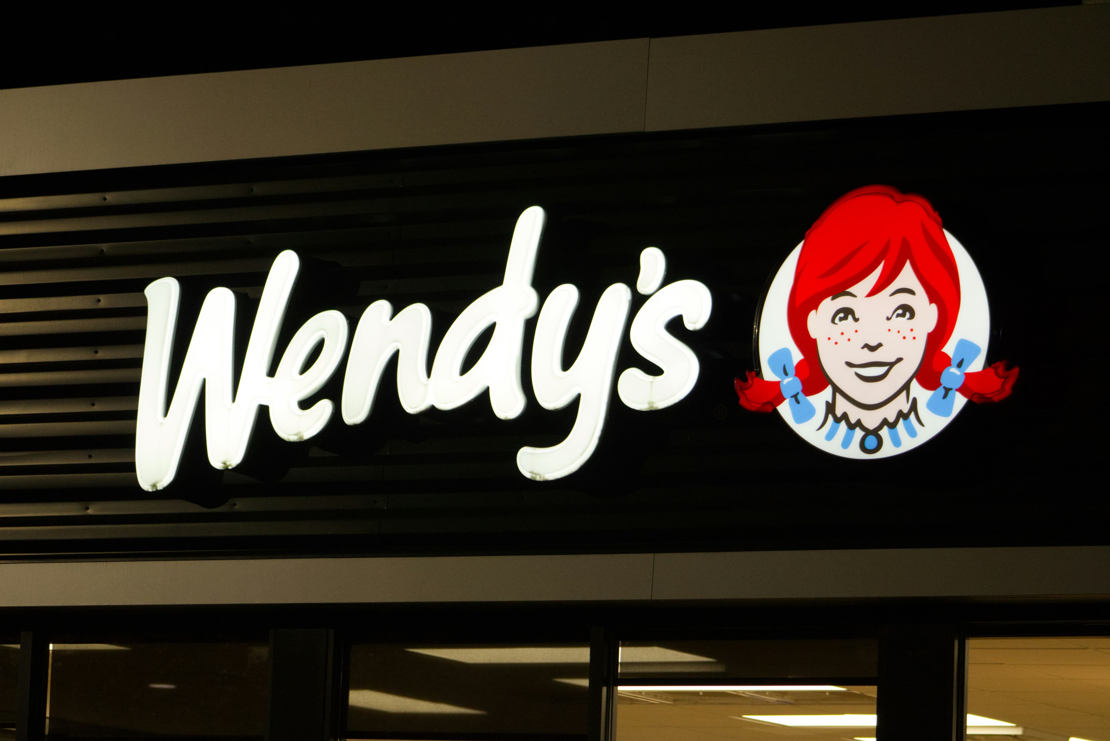 Illuminated Wendy's storefront sign at night, with the white script logo beside the red-haired mascot in blue pigtails on a black background.