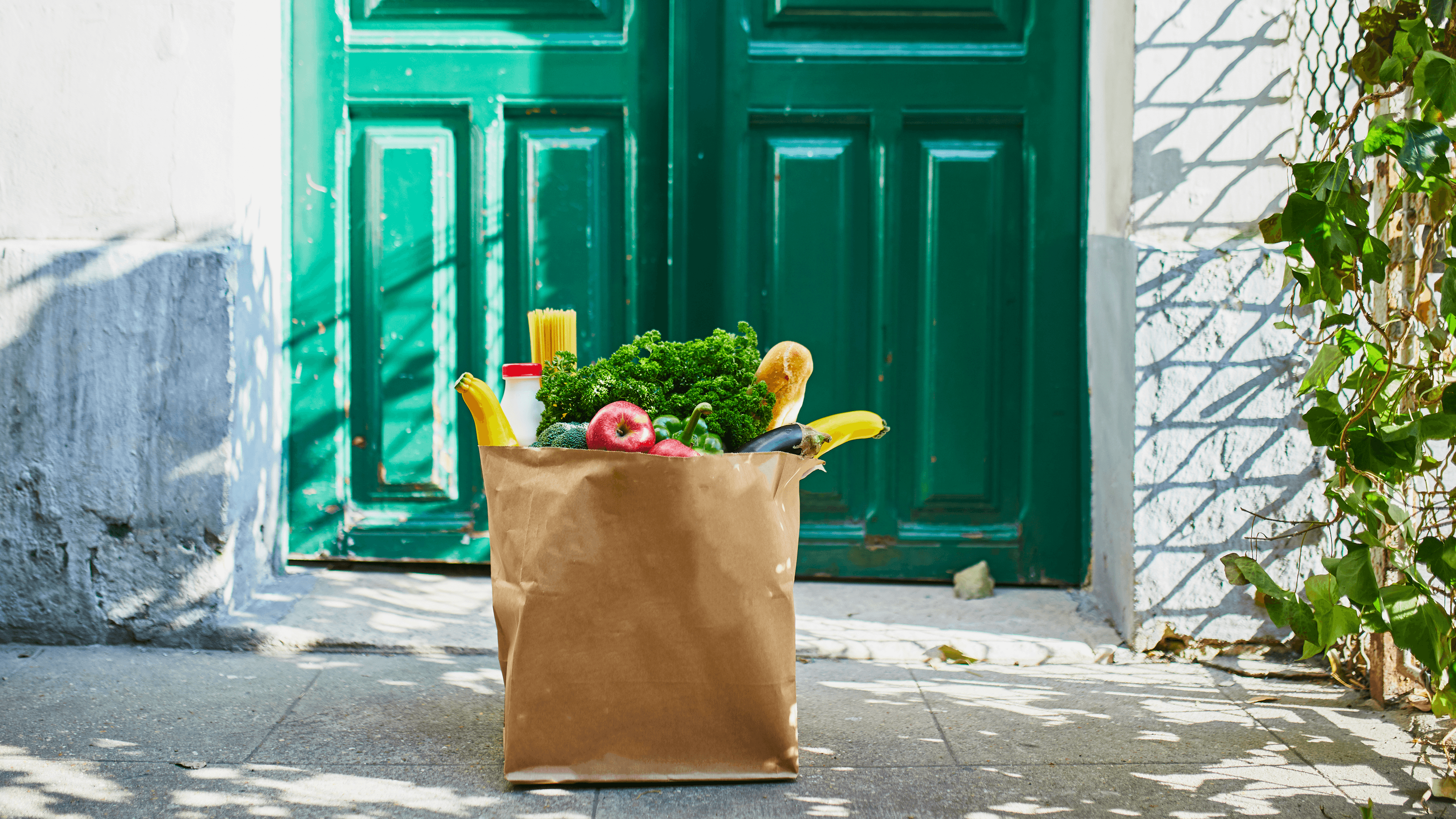 A brown paper grocery bag filled with fresh produce, including broccoli, apples, bananas, and greens, sitting on a concrete doorstep in front of a closed green front door.