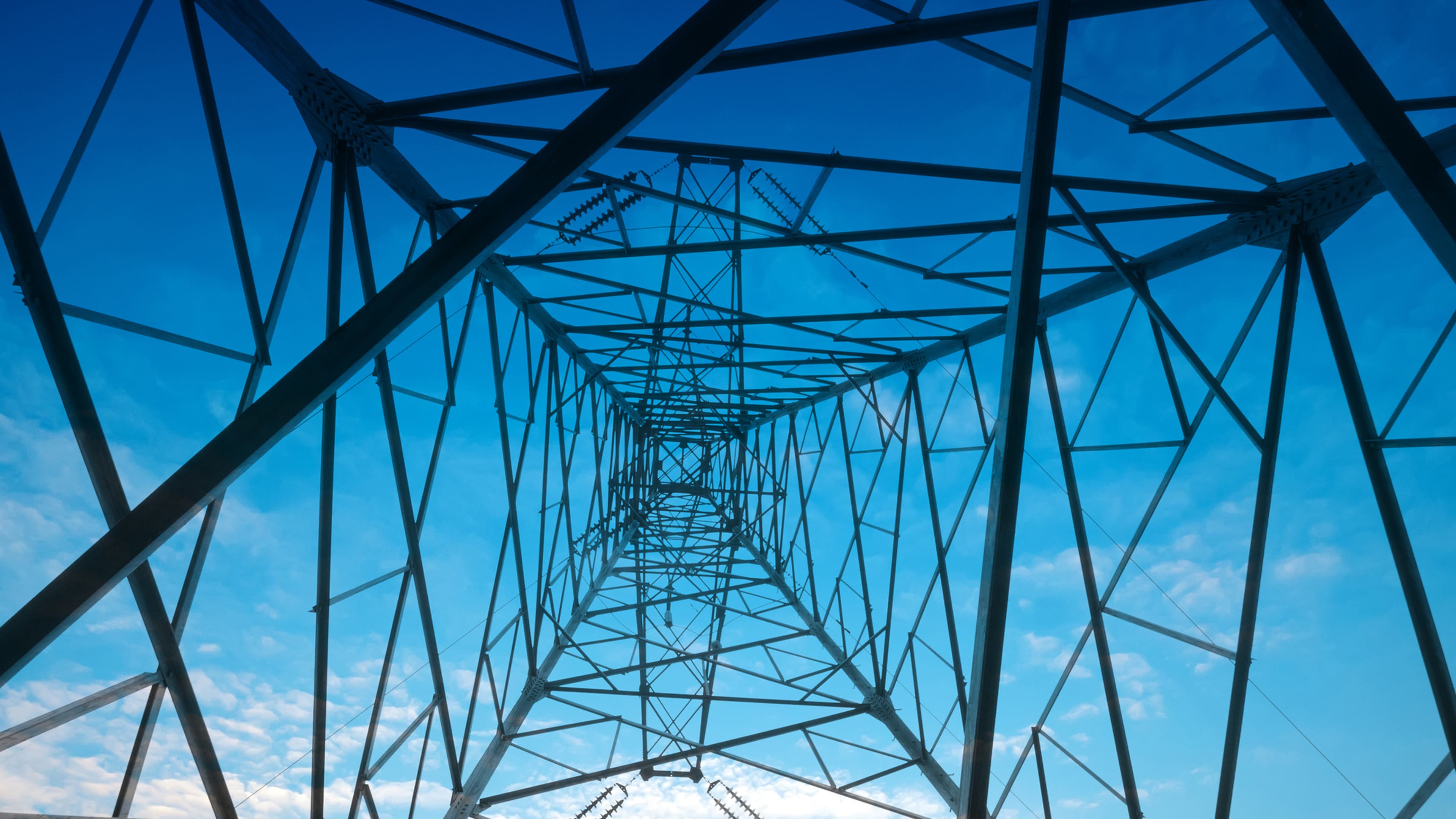 Looking directly up through the geometric steel lattice framework of a large power pylon against a blue sky.