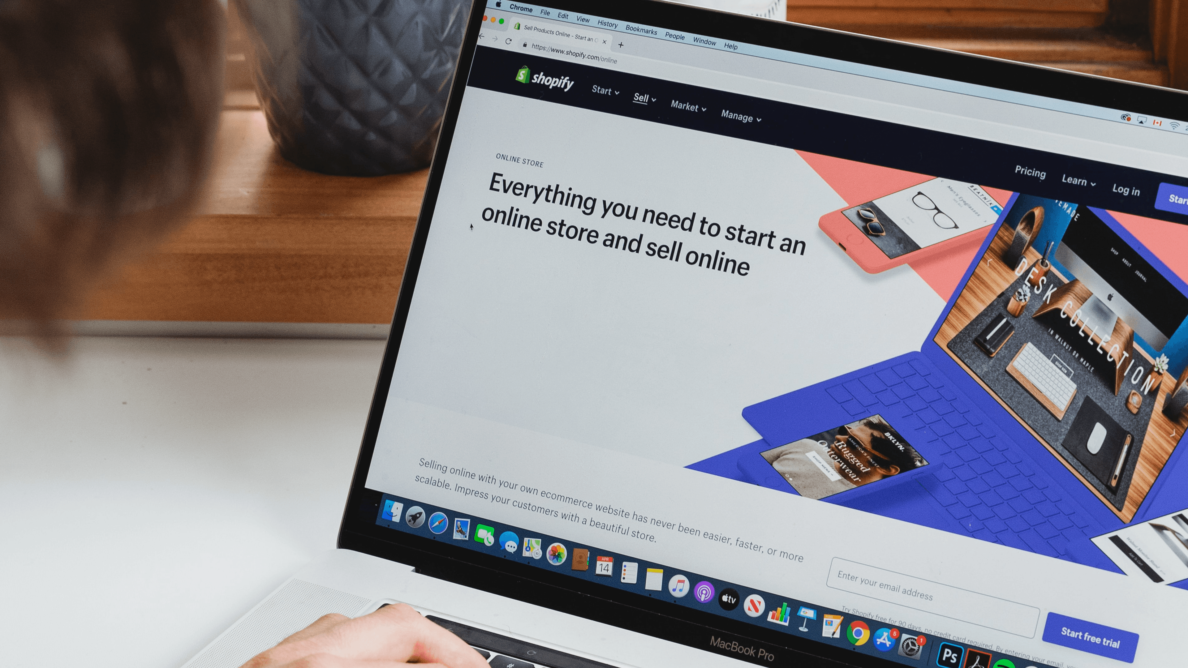 An over-the-shoulder view of a person using laptop with the screen displaying the official Shopify website homepage.